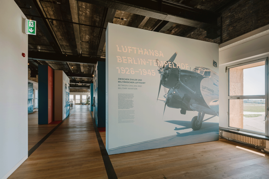 Lufthansa-Ausstellung mit großem Wandpanel zur Geschichte „Lufthansa Berlin‑Tempelhof 1926–1945“, darauf ein historisches Flugzeugfoto und zweisprachige Überschrift; im Hintergrund hohe Fenster mit Blick aufs Vorfeld sowie industriell anmutende Decke und Holzfußboden.