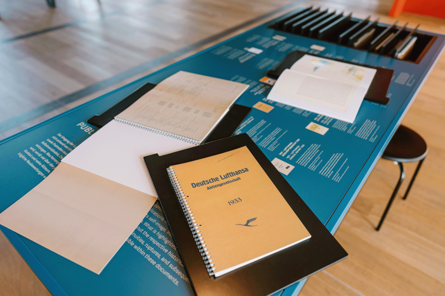 Ein Ausstellungstisch mit blauem Untergrund zeigt mehrere aufgeklappte und geschlossene spiralgebundene Hefte und Dokumente. Im Zentrum liegt ein gelbes Heft mit dem Titel „Deutsche Lufthansa Aktiengesellschaft 1933“ und Lufthansa‑Logo. Weitere Unterlagen mit Texten und Diagrammen sind darum angeordnet. Im Hintergrund stehen Stühle auf einem Holzfußboden, was auf eine Museums‑ oder Ausstellungssituation hinweist.