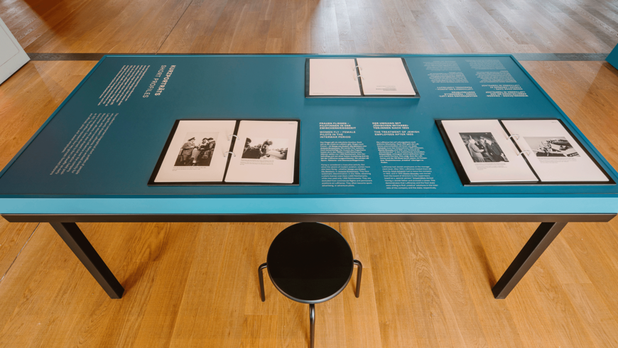Ein blauer Ausstellungstisch mit schwarzen Beinen zeigt mehrere aufgeklappte Ordner mit historischen Schwarz‑Weiß‑Fotografien und zweisprachigen Texten. Die sichtbaren Kapitelüberschriften befassen sich mit „Wartime Portraits“, „Frauen im Kriegseinsatz“ und „Der Umgang mit jüdischen Angestellten nach 1933“. Vor dem Tisch steht ein schwarzer Hocker, der Besucher*innen zum Lesen der Dokumente einlädt. Der Tisch steht auf einem hellen Holzfußboden in einer musealen Umgebung.