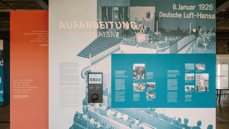 Eine Ausstellungswand im THF TOWER zeigt mehrere farblich abgesetzte Infotafeln zur Geschichte der Deutschen Luft‑Hansa. Links stehen orange, weiße und blaue Textmodule mit historischen Erläuterungen, Fotos und einem Audiostation‑Element mit Kopfhörern. Im Hintergrund ist ein großes Schwarz‑Weiß‑Foto einer historischen Veranstaltung mit sitzendem Publikum und einem Redner am Podium zu sehen. Überschriften thematisieren „Aufarbeitung“, „Kriegsverbrechen und personelle Kontinuitäten“ sowie „Traditionspflege und Firmenjubiläen“.