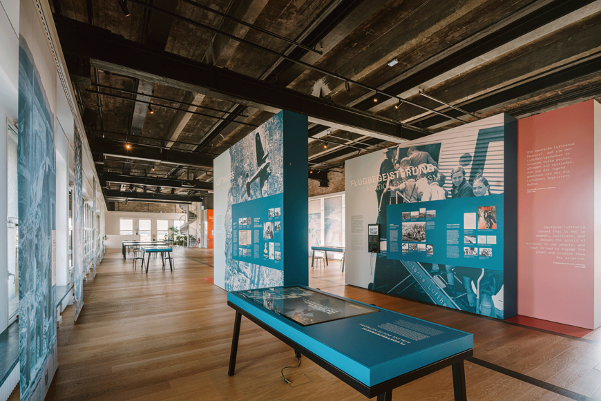 Ausstellungsraum im THF TOWER mit Holzfußboden, großen Informations- und Grafiktafeln zu Luftfahrtgeschichte (u. a. „Flugbegeisterung“ und „Lufthansa“) sowie Tischen und interaktiven Stationen; links fällt Tageslicht durch hohe Fenster in den Raum.